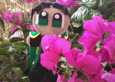 A close-up photo of a Loki-inspired felt plush doll in a garden with pink bougainvillea flowers.