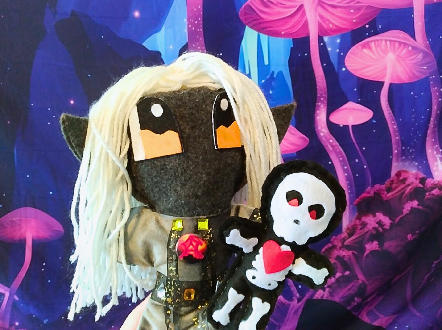 Photo of a dark elf wizard plush in a gem-lined gray robe, holding up smaller skeleton plush.