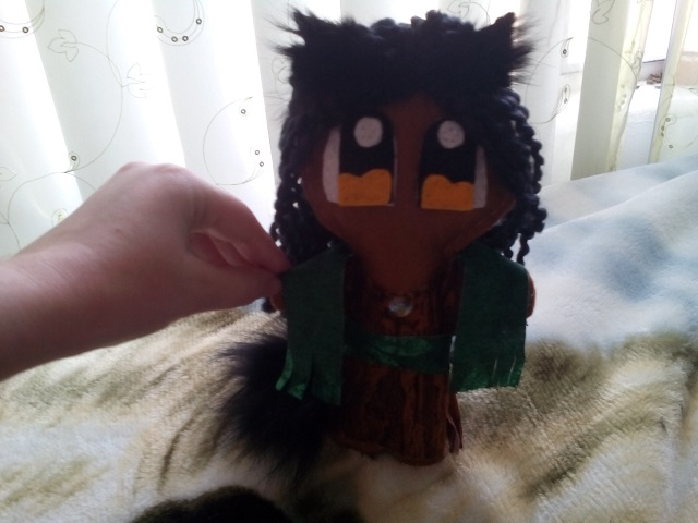 A small plush doll is held upright by an artist's hand. Plush is of a brown-skinned girl with black hair, furry ears, and a tail.