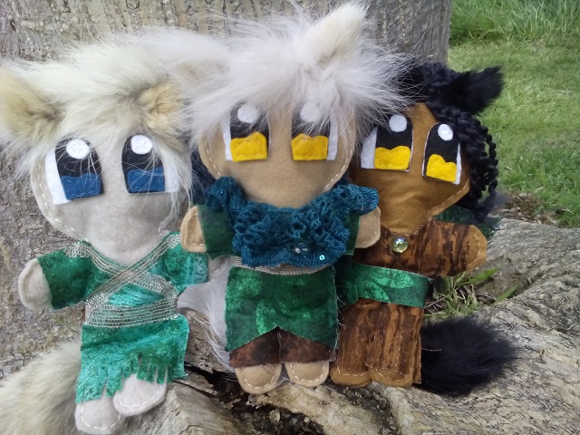 A trio of small plush dolls sitting on a tree trunk. One is wearing a teal dress, the other a green tunic, and the third wearing a brown tunic. All have fluffy ears and tails.