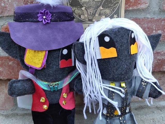 Two dark elf plush standing outside by a brick wall with a 'wanted' poster in the background. Wanted poster depicts one of the plush dolls.