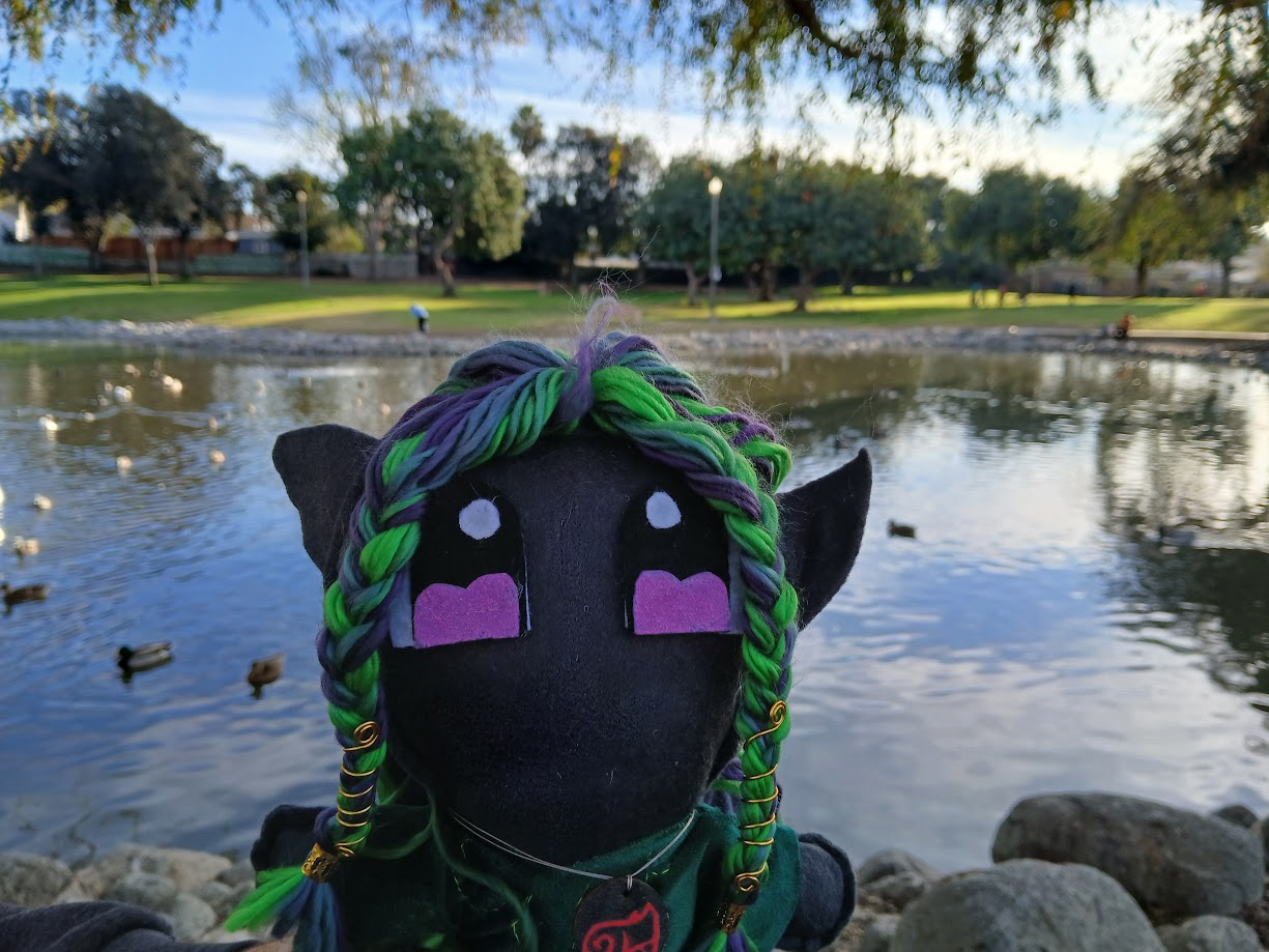 A plush doll featuring a dark elf with colorful green and purple braided hair stands in front of a duck pond in a park.