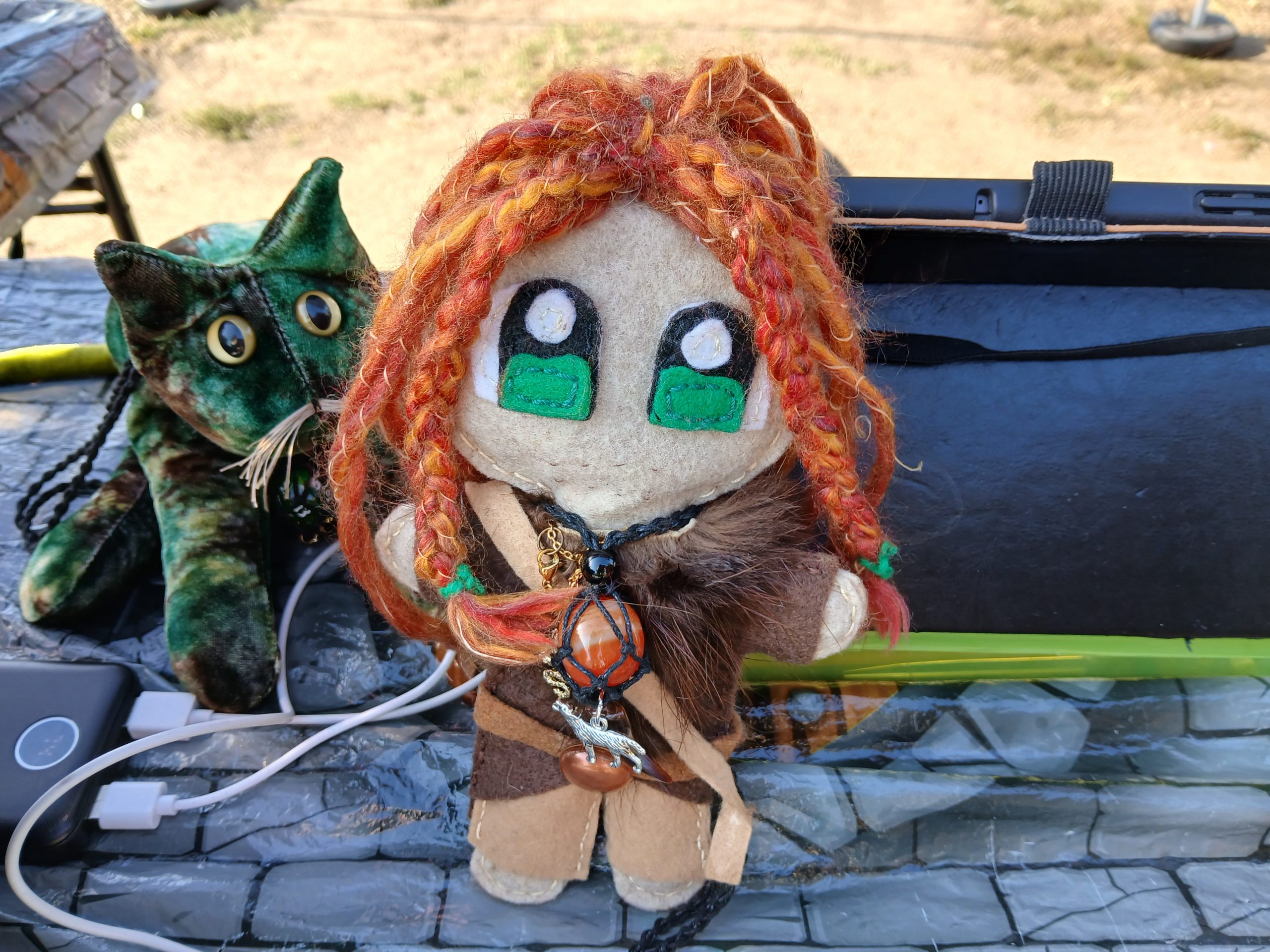 A slightly weathered plush doll with bright flame-red braided hair stands next to a green and gold cat plush on a market table.